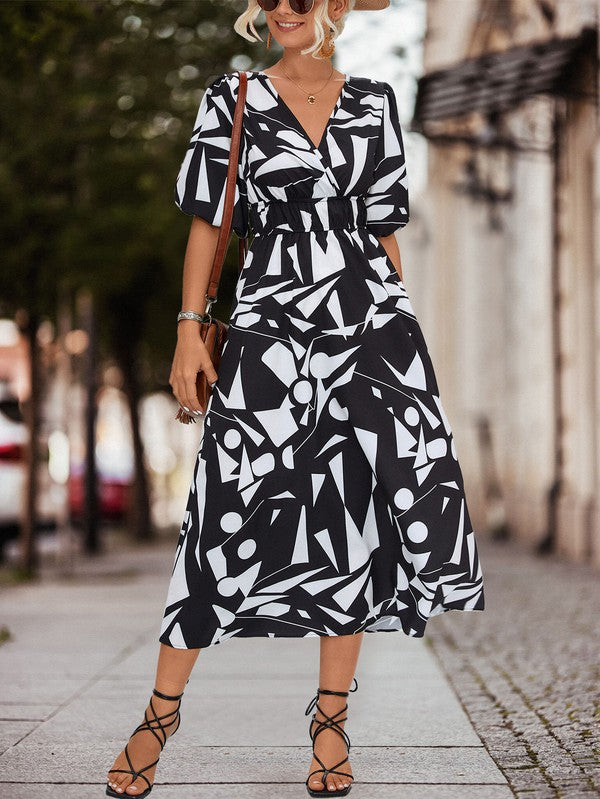 A person in a Women Printed Long Dress featuring a black and white geometric pattern and a V-neck stands outdoors, wearing strappy sandals and holding a brown handbag.