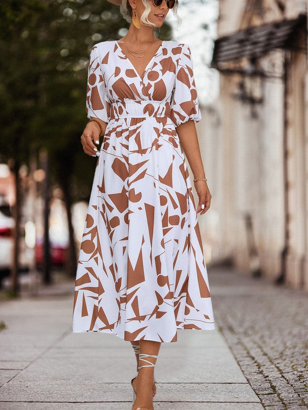 A person in a Women Printed Long Dress featuring a black and white geometric pattern and a V-neck stands outdoors, wearing strappy sandals and holding a brown handbag.