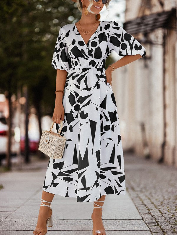 A person in a Women Printed Long Dress featuring a black and white geometric pattern and a V-neck stands outdoors, wearing strappy sandals and holding a brown handbag.