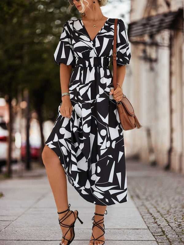 A person in a Women Printed Long Dress featuring a black and white geometric pattern and a V-neck stands outdoors, wearing strappy sandals and holding a brown handbag.