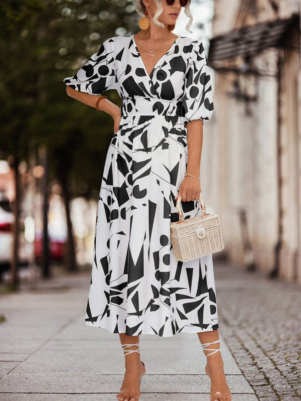 A person in a Women Printed Long Dress featuring a black and white geometric pattern and a V-neck stands outdoors, wearing strappy sandals and holding a brown handbag.