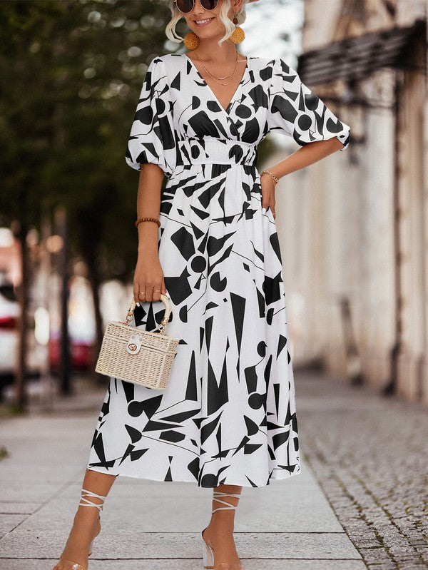 A person in a Women Printed Long Dress featuring a black and white geometric pattern and a V-neck stands outdoors, wearing strappy sandals and holding a brown handbag.