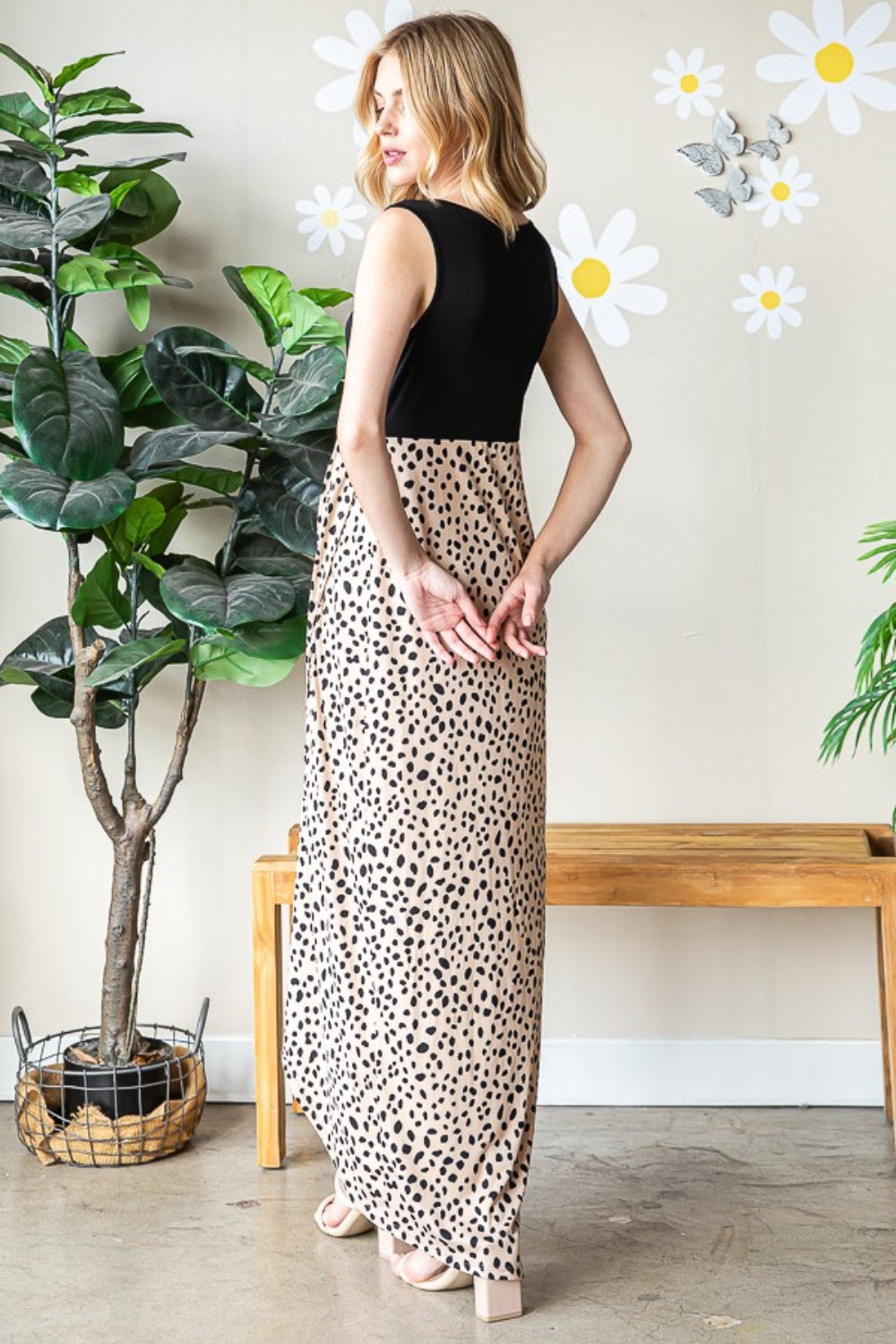 A woman poses indoors wearing the Heimish Full Size Slit Animal Print V-Neck Wide Strap Dress. With one hand resting on her head, she stands elegantly surrounded by potted plants and wall decorations.