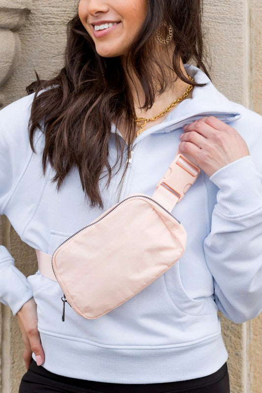 A woman stands outdoors wearing a white button-up shirt, blue jeans, and a Roam Nylon Belt Sling Bag with an adjustable strap. She has long brown hair and is smiling slightly.