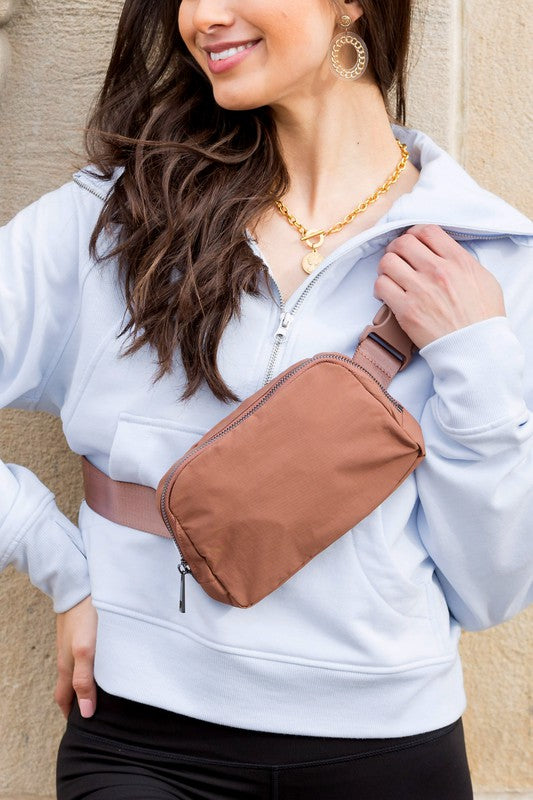 A woman stands outdoors wearing a white button-up shirt, blue jeans, and a Roam Nylon Belt Sling Bag with an adjustable strap. She has long brown hair and is smiling slightly.