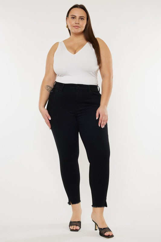 A person poses against a plain white background wearing a white sleeveless top, Plus High Rise Ankle Skinny Jeans featuring a frayed hem, and black heels. They have long, straight hair and display a visible tattoo on their left upper arm.