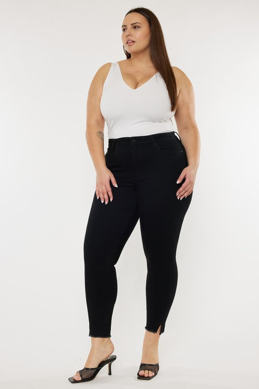 A person poses against a plain white background wearing a white sleeveless top, Plus High Rise Ankle Skinny Jeans featuring a frayed hem, and black heels. They have long, straight hair and display a visible tattoo on their left upper arm.