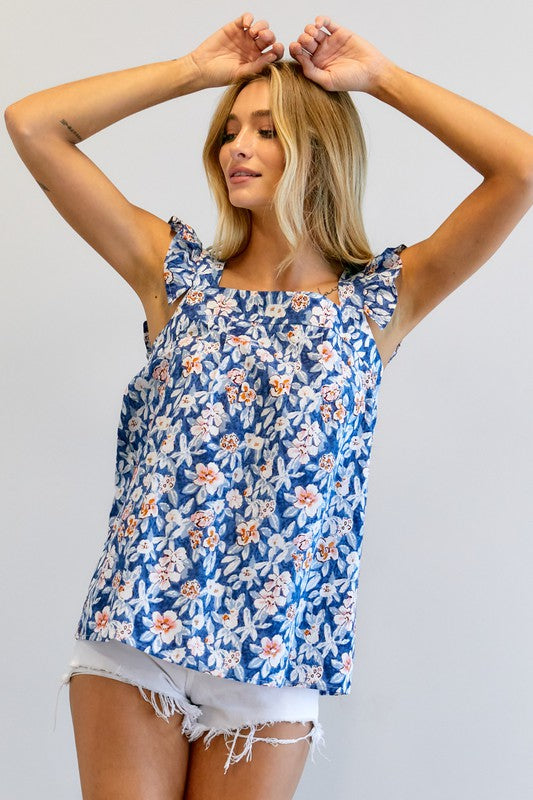 A woman in a vibrant summer wardrobe stands against a plain background, wearing the Floral Printed Ruffle Sleeveless Top paired with white shorts, with one hand casually tucked in her pocket.