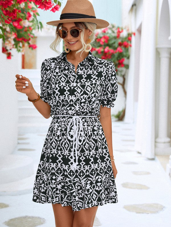 Dressed in a Women Print Button Down Jumpsuit, a woman accessorized with a stylish hat and sunglasses holds a beige purse. She stands against the backdrop of a pristine white building, framed by vibrant red flowers.