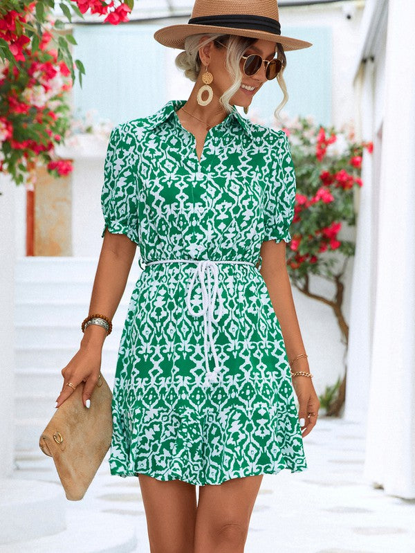 Dressed in a Women Print Button Down Jumpsuit, a woman accessorized with a stylish hat and sunglasses holds a beige purse. She stands against the backdrop of a pristine white building, framed by vibrant red flowers.