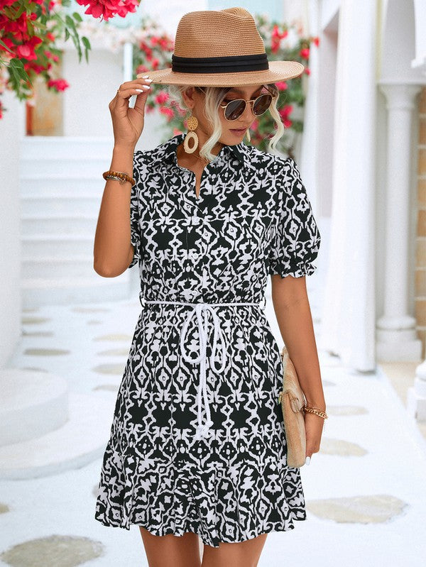 Dressed in a Women Print Button Down Jumpsuit, a woman accessorized with a stylish hat and sunglasses holds a beige purse. She stands against the backdrop of a pristine white building, framed by vibrant red flowers.