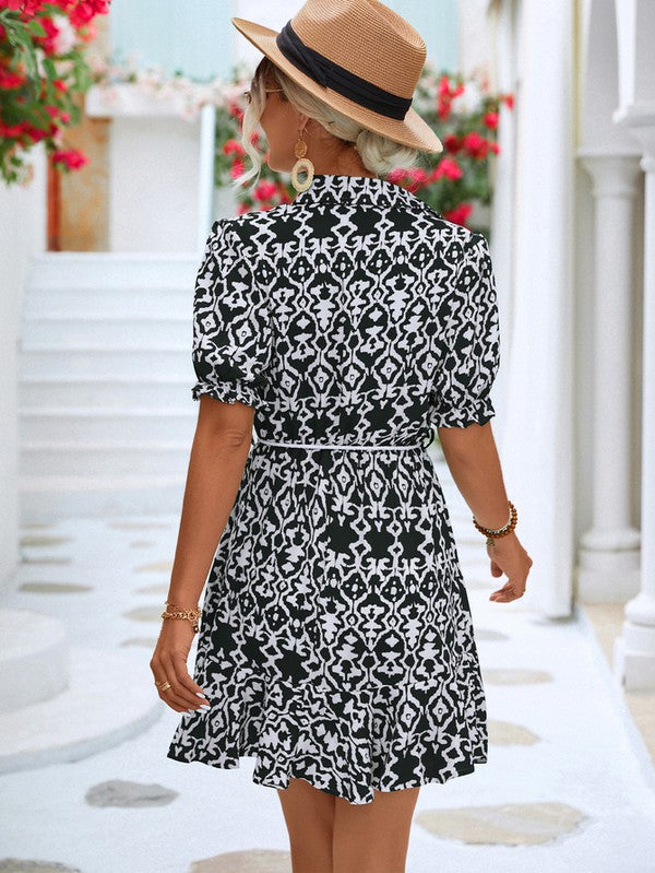 Dressed in a Women Print Button Down Jumpsuit, a woman accessorized with a stylish hat and sunglasses holds a beige purse. She stands against the backdrop of a pristine white building, framed by vibrant red flowers.