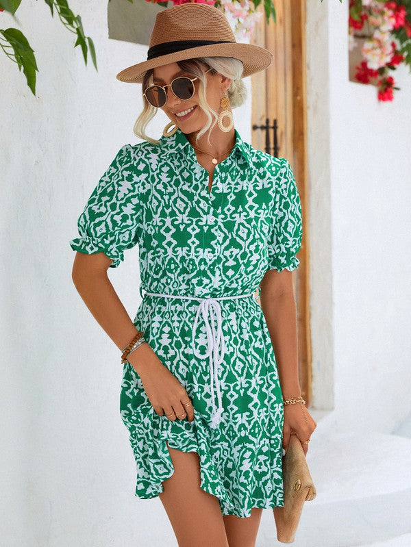 Dressed in a Women Print Button Down Jumpsuit, a woman accessorized with a stylish hat and sunglasses holds a beige purse. She stands against the backdrop of a pristine white building, framed by vibrant red flowers.