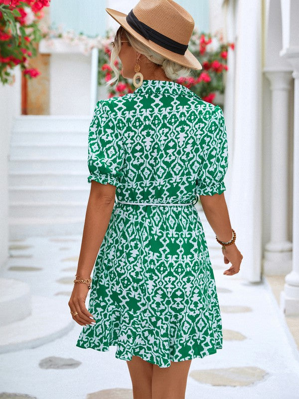 Dressed in a Women Print Button Down Jumpsuit, a woman accessorized with a stylish hat and sunglasses holds a beige purse. She stands against the backdrop of a pristine white building, framed by vibrant red flowers.