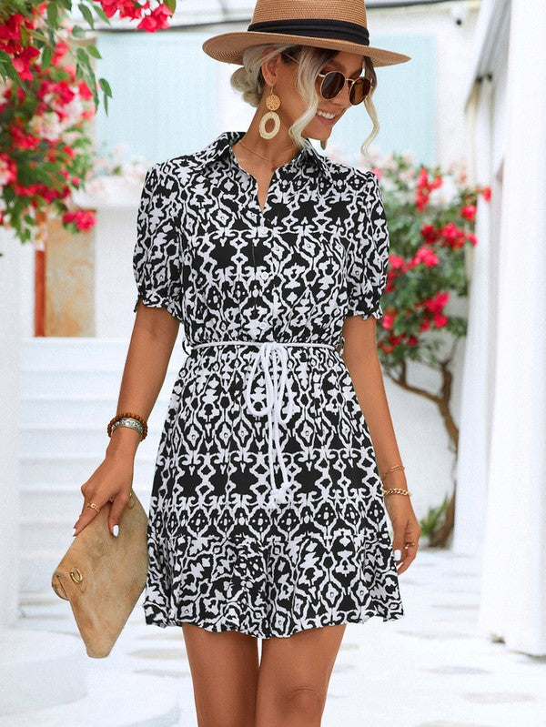 Dressed in a Women Print Button Down Jumpsuit, a woman accessorized with a stylish hat and sunglasses holds a beige purse. She stands against the backdrop of a pristine white building, framed by vibrant red flowers.