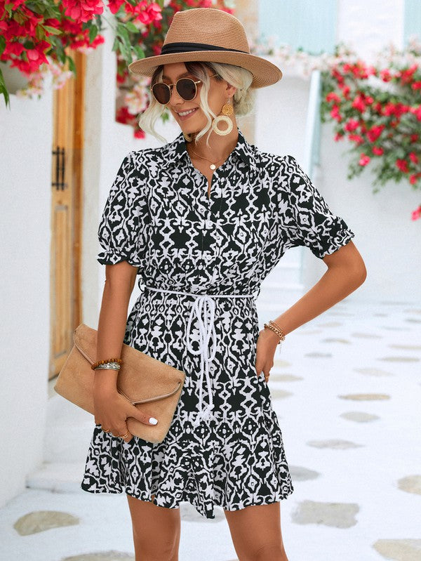 Dressed in a Women Print Button Down Jumpsuit, a woman accessorized with a stylish hat and sunglasses holds a beige purse. She stands against the backdrop of a pristine white building, framed by vibrant red flowers.