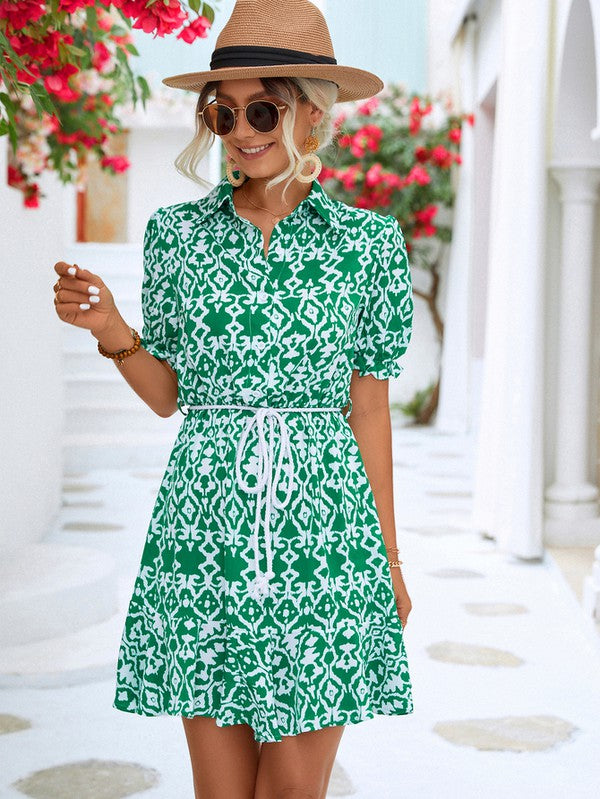 Dressed in a Women Print Button Down Jumpsuit, a woman accessorized with a stylish hat and sunglasses holds a beige purse. She stands against the backdrop of a pristine white building, framed by vibrant red flowers.