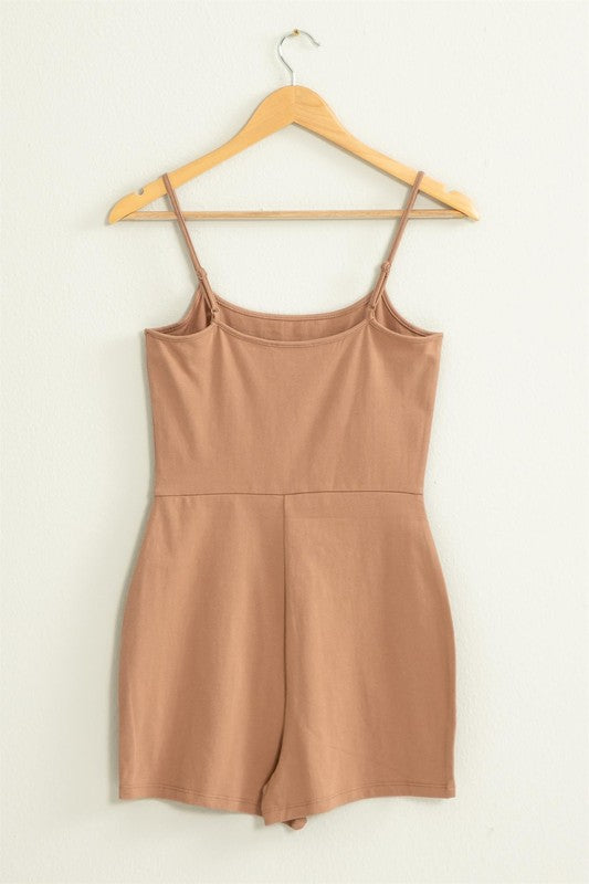 The Loving Me Fitted Sleeveless Romper, featuring a fitted bodice and spaghetti straps in black, hangs gracefully on a wooden hanger against a white wall.