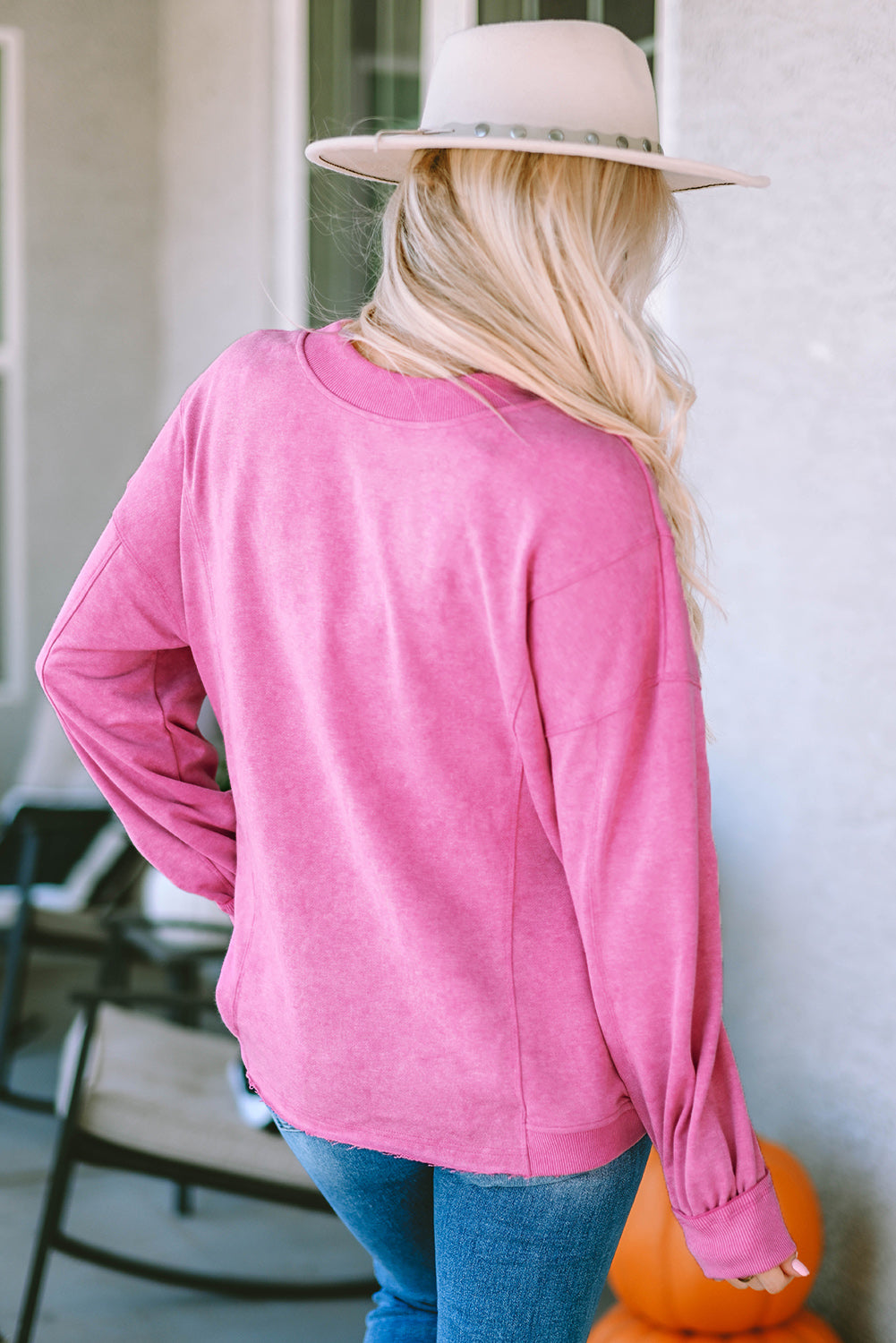 A woman stands with her back to the camera, wearing a long-sleeved Rose Exposed Seam Twist Open Back Oversized Sweatshirt and light blue jeans, showcasing a relaxed fit.