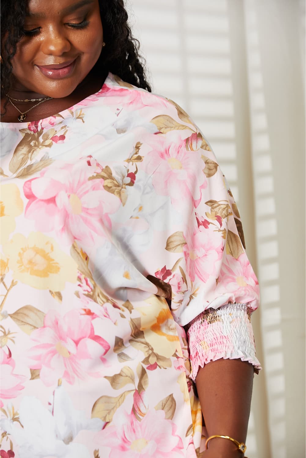 A woman in a Double Take Floral Round Neck Three-Quarter Sleeve Top and jeans stands smiling in a brightly lit room with a white background and drawers on the side.