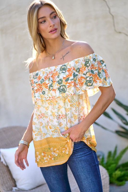 A woman with blonde hair is smiling while standing outdoors, wearing the Printed Off Shoulder Smocked Top paired with jeans.