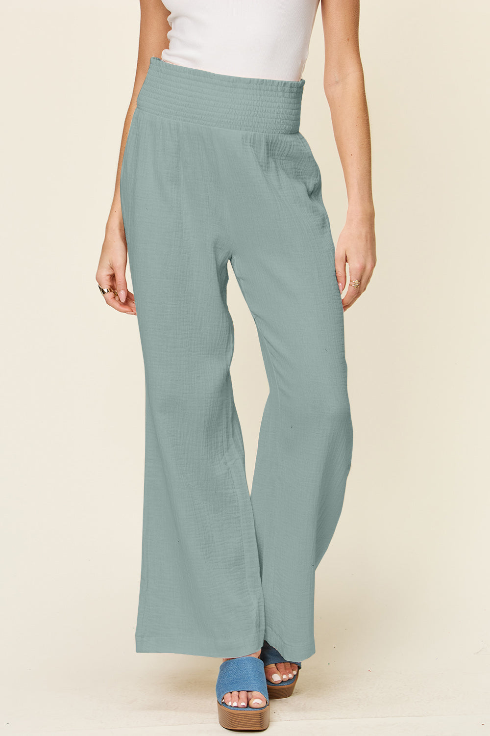 A person wearing Double Take Full Size Texture Smocked Waist Wide Leg Pants in black and a white 100% cotton top is standing with one hand on their hip. They are also wearing blue open-toe sandals.