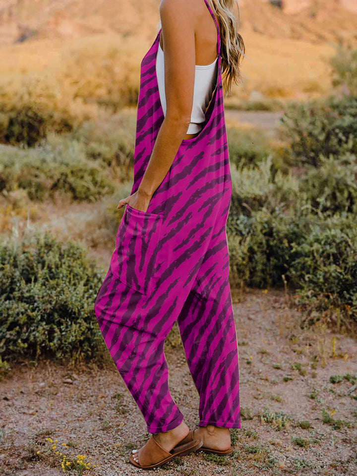 A person wearing a casual white V-neck tank top and a Full Size Printed V-Neck Sleeveless Jumpsuit with a brown animal print zebra-stripe pattern stands outdoors with one hand on their hat and the other in their pocket, set against a rocky landscape backdrop.