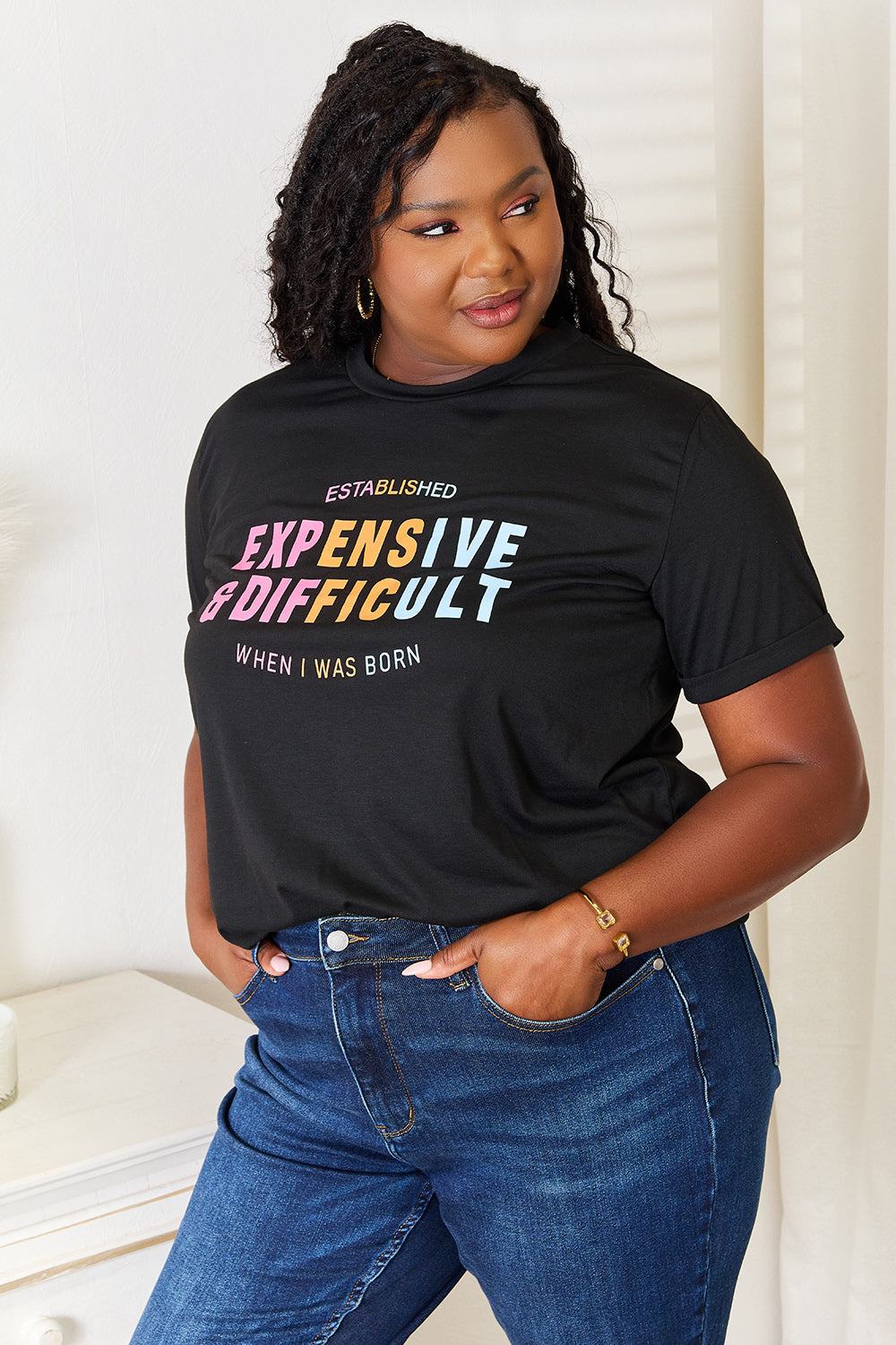 A woman with long braided hair, wearing the Simply Love Slogan Graphic Cuffed Sleeve T-Shirt that says "Established Expensive & Difficult When I Was Born," stands with one hand in her pocket. The trendy cuffed sleeves of her shirt add an extra touch of style.