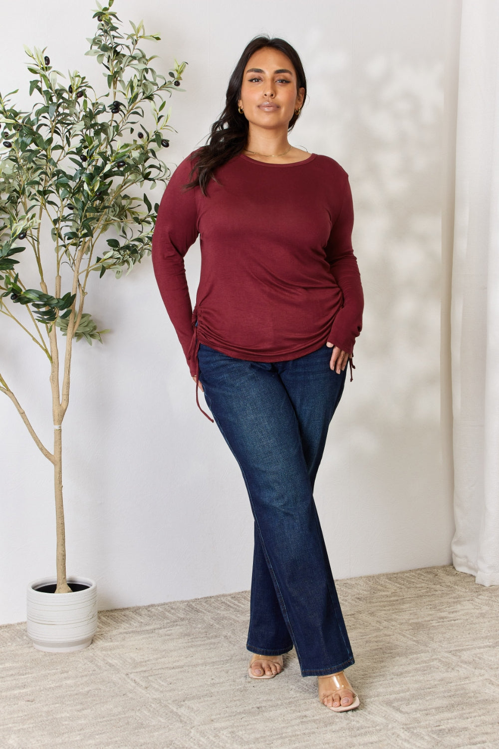 A woman stands in front of a plain background and a small tree, wearing the Culture Code Full Size Drawstring Round Neck Long Sleeve Top in maroon and blue jeans.