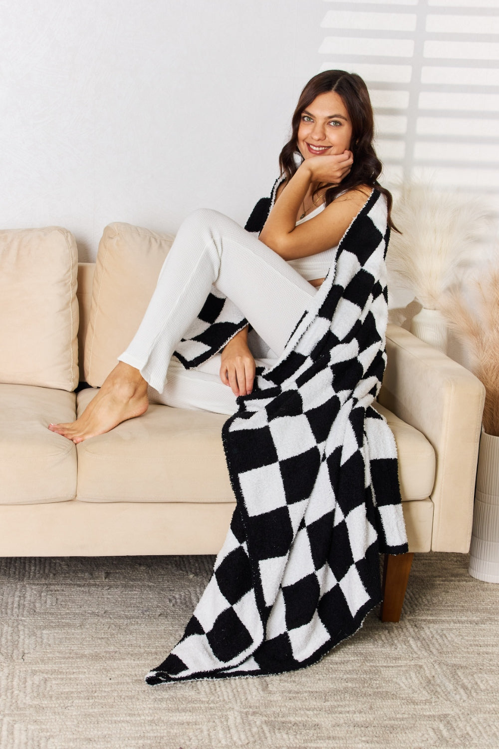 A person stands holding a large Cuddley Checkered Decorative Throw Blanket, smiling, in a room with a sofa and a potted plant. The blanket features a black and white checkerboard pattern, is made from 100% polyester, and imported for quality.