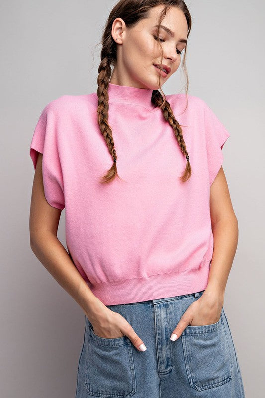 A person stands with hands in blue denim jeans pockets, wearing a pink Mock Neck Short Sleeve Top—a versatile wardrobe staple. The person has two braided pigtails and is looking slightly downward.