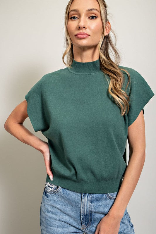 A person stands with hands in blue denim jeans pockets, wearing a pink Mock Neck Short Sleeve Top—a versatile wardrobe staple. The person has two braided pigtails and is looking slightly downward.