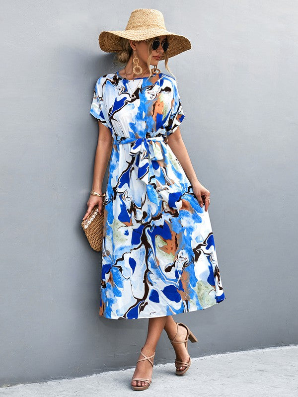 A woman in a Women Print Round Neck Short Sleeve Dress, complemented by a straw hat and sunglasses, gracefully strolls across a gray textured surface.