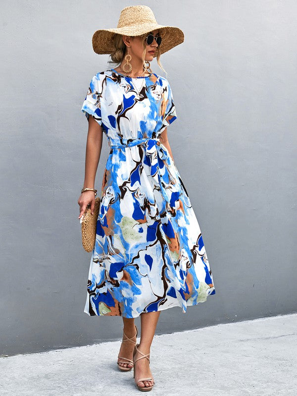 A woman in a Women Print Round Neck Short Sleeve Dress, complemented by a straw hat and sunglasses, gracefully strolls across a gray textured surface.