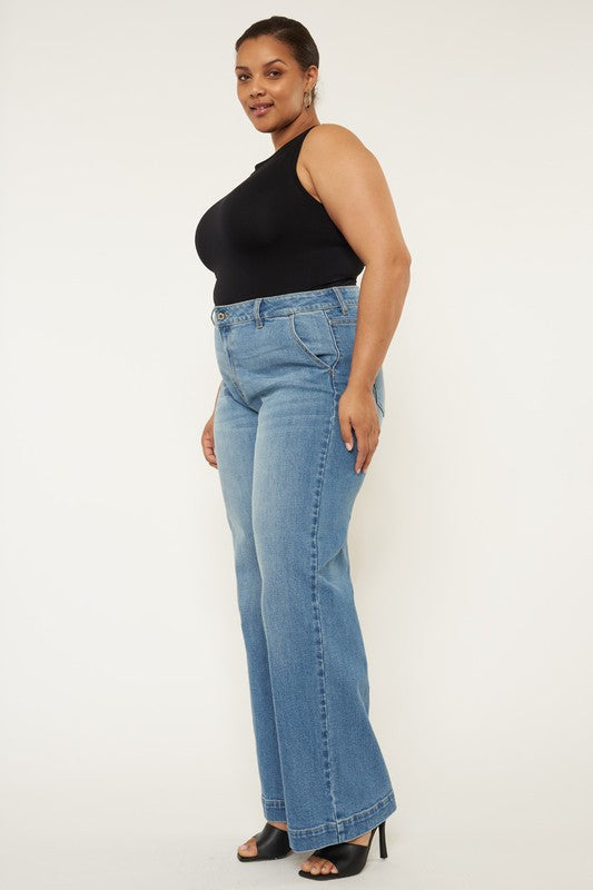 A person stands in a neutral pose, wearing a black sleeveless top, medium stone wash *Plus High Rise Trouser Wide Leg Jeans* made from comfort stretch denim, and black open-toe shoes, against a plain white background.