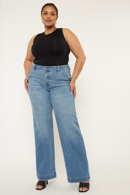 A person stands in a neutral pose, wearing a black sleeveless top, medium stone wash *Plus High Rise Trouser Wide Leg Jeans* made from comfort stretch denim, and black open-toe shoes, against a plain white background.