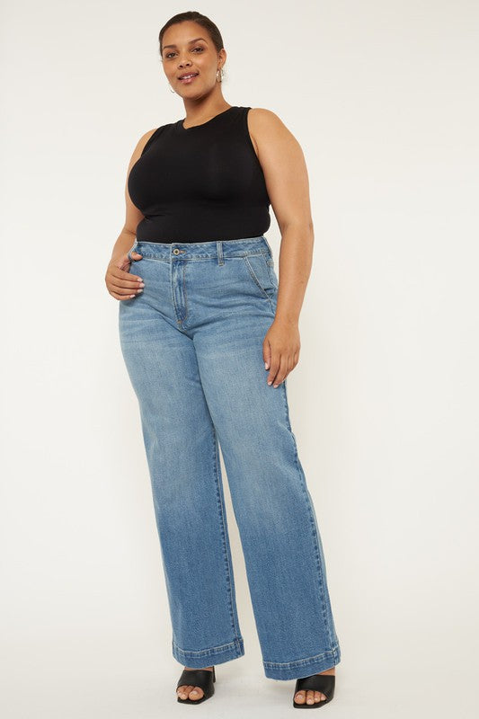 A person stands in a neutral pose, wearing a black sleeveless top, medium stone wash *Plus High Rise Trouser Wide Leg Jeans* made from comfort stretch denim, and black open-toe shoes, against a plain white background.