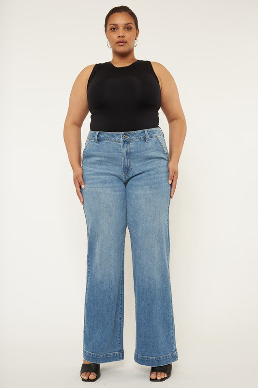 A person stands in a neutral pose, wearing a black sleeveless top, medium stone wash *Plus High Rise Trouser Wide Leg Jeans* made from comfort stretch denim, and black open-toe shoes, against a plain white background.