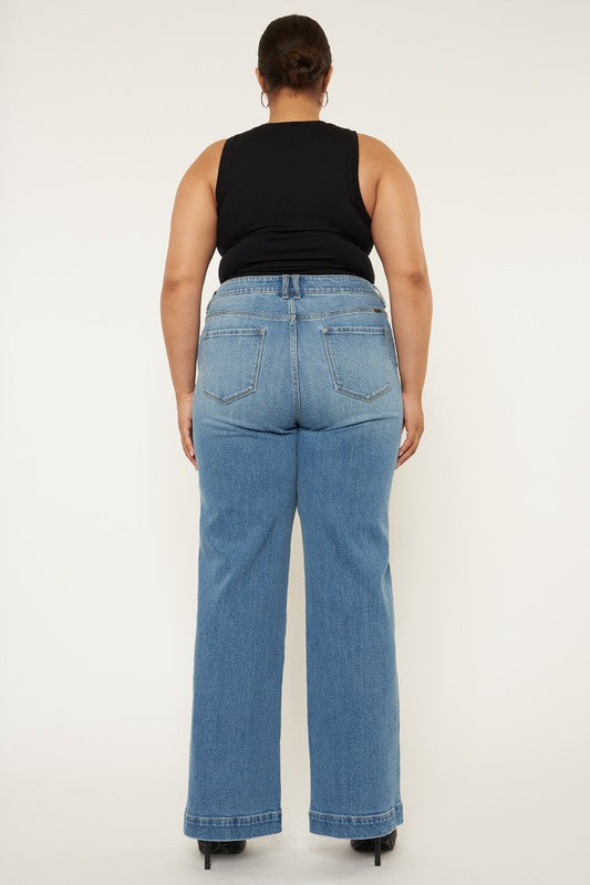 A person stands in a neutral pose, wearing a black sleeveless top, medium stone wash *Plus High Rise Trouser Wide Leg Jeans* made from comfort stretch denim, and black open-toe shoes, against a plain white background.