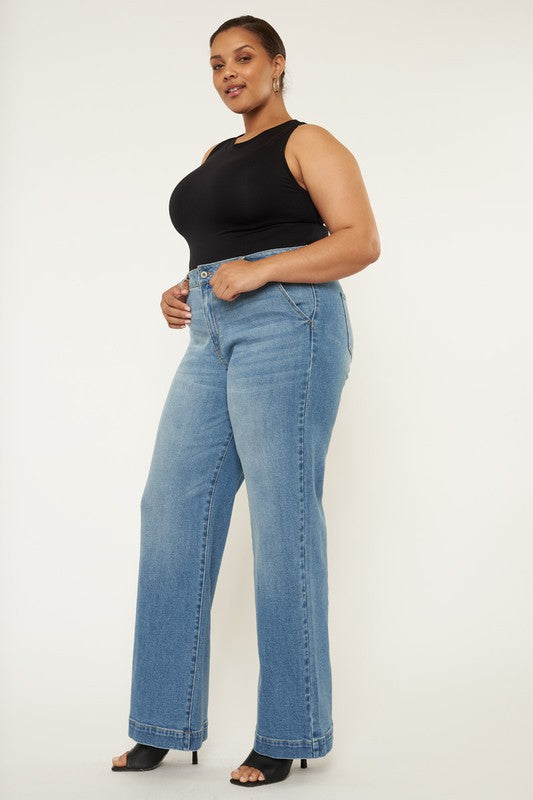 A person stands in a neutral pose, wearing a black sleeveless top, medium stone wash *Plus High Rise Trouser Wide Leg Jeans* made from comfort stretch denim, and black open-toe shoes, against a plain white background.