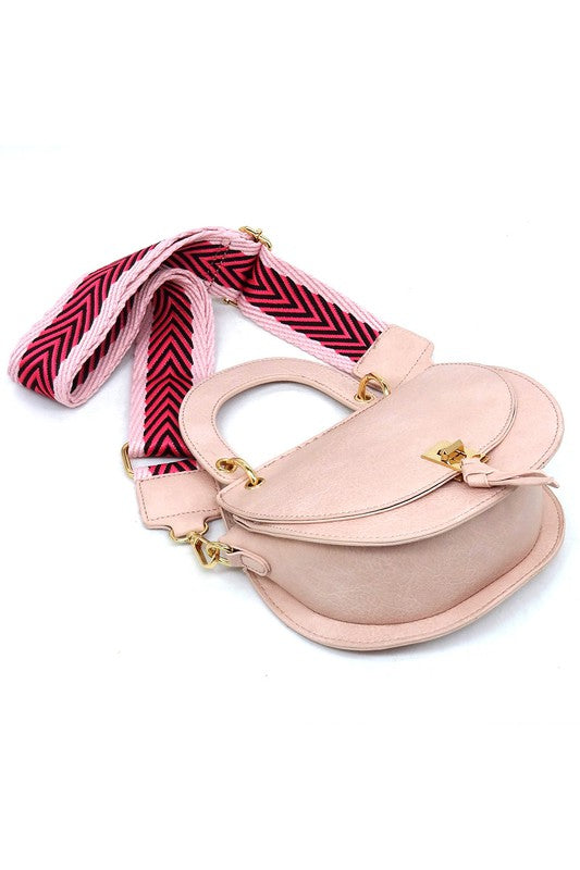 The Fashion Flap Saddle Satchel Crossbody Bag is a light pink handbag crafted from faux vegan leather and adorned with gold hardware. It features a buckle clasp and comes with a detachable, wide strap that has a red and black chevron pattern with pink trim, allowing it to double as a chic crossbody bag.