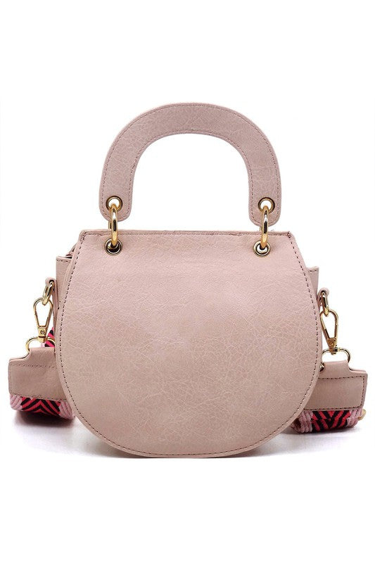 The Fashion Flap Saddle Satchel Crossbody Bag is a light pink handbag crafted from faux vegan leather and adorned with gold hardware. It features a buckle clasp and comes with a detachable, wide strap that has a red and black chevron pattern with pink trim, allowing it to double as a chic crossbody bag.