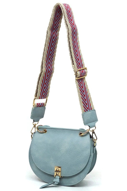 The Fashion Flap Saddle Satchel Crossbody Bag is a light pink handbag crafted from faux vegan leather and adorned with gold hardware. It features a buckle clasp and comes with a detachable, wide strap that has a red and black chevron pattern with pink trim, allowing it to double as a chic crossbody bag.