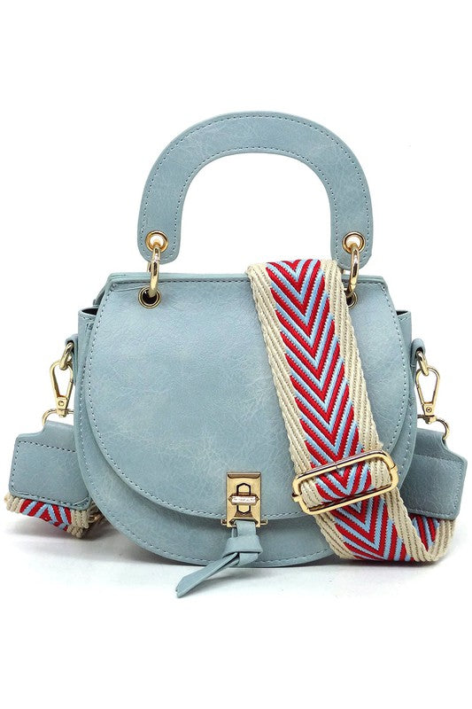 The Fashion Flap Saddle Satchel Crossbody Bag is a light pink handbag crafted from faux vegan leather and adorned with gold hardware. It features a buckle clasp and comes with a detachable, wide strap that has a red and black chevron pattern with pink trim, allowing it to double as a chic crossbody bag.
