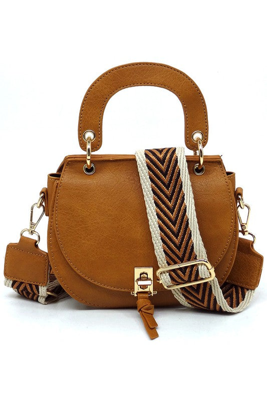 The Fashion Flap Saddle Satchel Crossbody Bag is a light pink handbag crafted from faux vegan leather and adorned with gold hardware. It features a buckle clasp and comes with a detachable, wide strap that has a red and black chevron pattern with pink trim, allowing it to double as a chic crossbody bag.