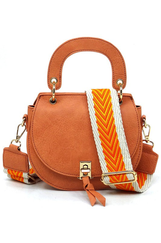The Fashion Flap Saddle Satchel Crossbody Bag is a light pink handbag crafted from faux vegan leather and adorned with gold hardware. It features a buckle clasp and comes with a detachable, wide strap that has a red and black chevron pattern with pink trim, allowing it to double as a chic crossbody bag.