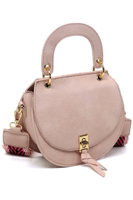 The Fashion Flap Saddle Satchel Crossbody Bag is a light pink handbag crafted from faux vegan leather and adorned with gold hardware. It features a buckle clasp and comes with a detachable, wide strap that has a red and black chevron pattern with pink trim, allowing it to double as a chic crossbody bag.