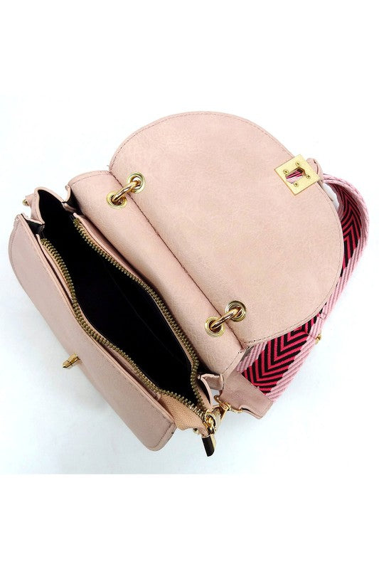 The Fashion Flap Saddle Satchel Crossbody Bag is a light pink handbag crafted from faux vegan leather and adorned with gold hardware. It features a buckle clasp and comes with a detachable, wide strap that has a red and black chevron pattern with pink trim, allowing it to double as a chic crossbody bag.