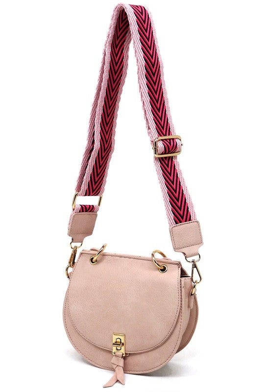 The Fashion Flap Saddle Satchel Crossbody Bag is a light pink handbag crafted from faux vegan leather and adorned with gold hardware. It features a buckle clasp and comes with a detachable, wide strap that has a red and black chevron pattern with pink trim, allowing it to double as a chic crossbody bag.