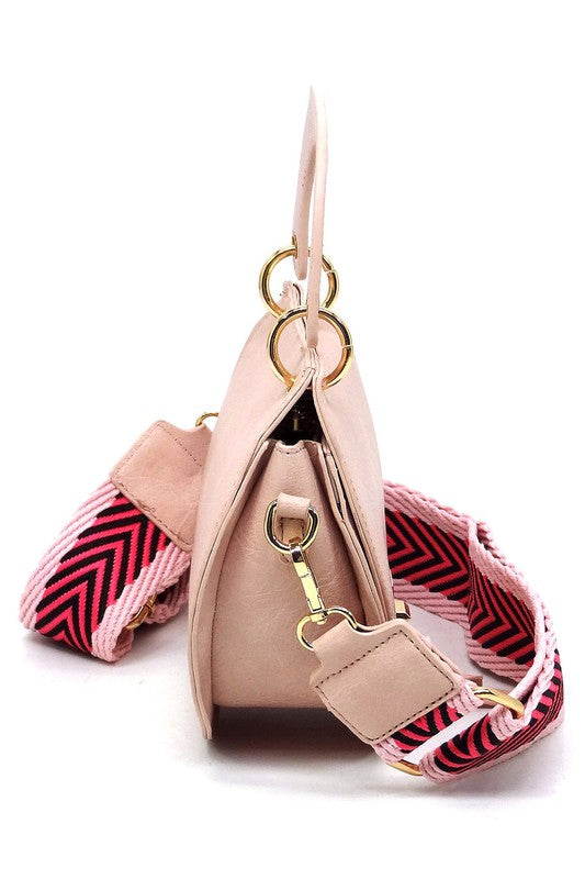 The Fashion Flap Saddle Satchel Crossbody Bag is a light pink handbag crafted from faux vegan leather and adorned with gold hardware. It features a buckle clasp and comes with a detachable, wide strap that has a red and black chevron pattern with pink trim, allowing it to double as a chic crossbody bag.